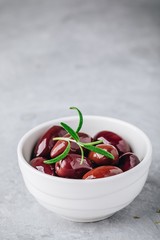 Fresh black olives in olive oil in white bowl with rosemary on gray stone background.