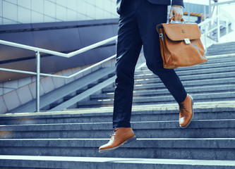 Businessman making steps down stairs. Cropped shot of man wearing elegant suit and shoes walking down stairs and holding brown leather bag in his hand.