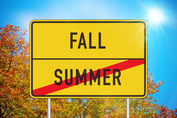 Yellow roadsign with fall and summer written on it against blue sky and autumn trees in background
