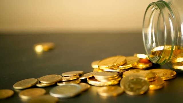 Yellow coins fell out from jar. Symbol of investing, keeping money concept. Collecting cash conis in glass tin as moneybox. Close-up still life with gold coins on black table and rotating penny.