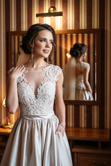 Portrait of a beautiful bride in a wedding dress standing in a room near a mirror. Vertical frame