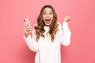 Photo of happy young woman with beautiful long hair screaming and clenching fist while holding mobile phone in hand, isolated over pink background