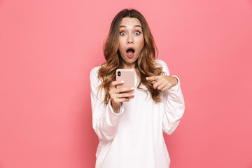 Image of excited caucasian woman with beautiful long hair screaming while holding cell phone in hand, isolated over pink background