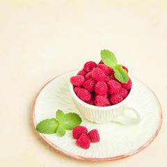 Ripe raspberries with green mint leaves in cup and saucer on pastel yellow background.