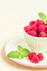 Ripe raspberries with green mint leaves in cup and saucer on pastel yellow background.