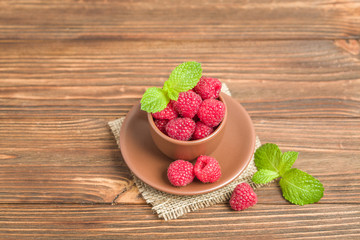 Ripe raspberries with green mint leaves in brown cup and saucer on sackcloth and wooden background.