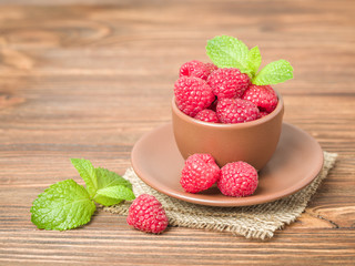 Ripe raspberries with green mint leaves in brown cup and saucer on sackcloth and wooden background.