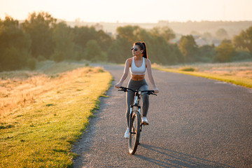 Beautiful happy fit and sporty woman in sportswear riding bicycle on the countryside road near the field on sunset.