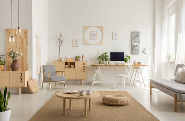 Fototapeta na wymiar Wooden table and pouf on carpet in white living room interior with poster and workspace. Real photo