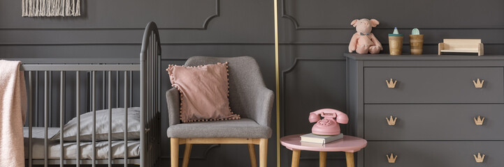 Modern gray armchair by an elegant wall with wainscoting, between a classic, wooden baby crib and a scandi dresser in a dark nursery bedroom interior