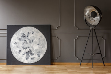 Big handmade moon painting placed on wooden floor in real photo of dark room interior with wainscoting wall and metal studio lamp