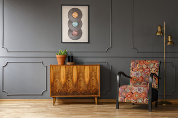 Real photo of wooden retro cupboard with fresh plant and candle standing in dark grey living room interior with colorful armchair, gold lamp and poster with vinyls