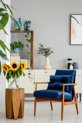 Sunflowers on wooden stool next to blue armchair in living room interior with poster. Real photo