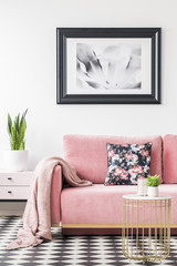 Floral pillows and blanket on pink couch in living room interior with poster and plant. Real photo