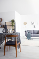 Table and wooden chair in white apartment interior with plant and grey couch with blanket. Real photo