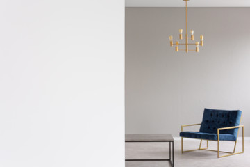 Copy space on white empty wall in flat interior with gold chandelier above blue armchair in blurred...