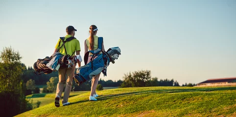 Poster Im Rahmen In voller Länge eines glücklichen Paares mit einem gesunden Lebensstil, das Golf-Outfits trägt, während es an einem sonnigen Sommertag Standbags mit professionellen Schlägern zum Golfplatz trägt © Kzenon