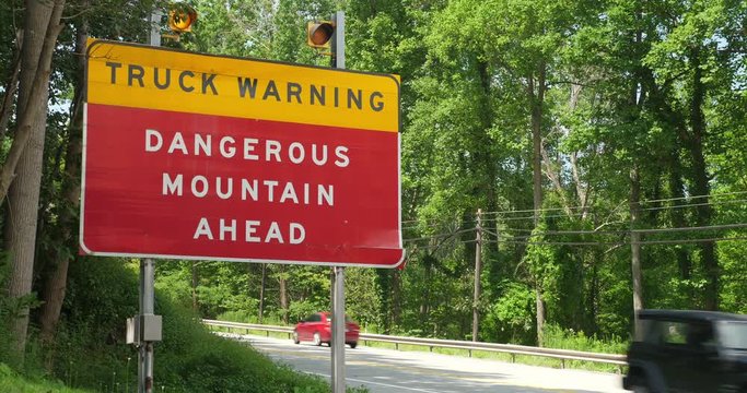 A large red sign on a mountainous Pennsylvania road warning trucks of a long downward slope ahead. Traffic passes behind, flashing yellow lights atop.  	