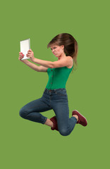 Image of young woman over green background using laptop computer or tablet gadget while jumping.