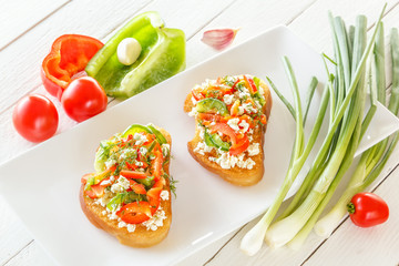 Two Bruschetta with red sweet pepper and goat cheese in a plate next to fresh vegetables on white boards