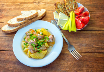 Served lunch or dinner – stewed potato with meat and cut green onion, pepper, tomato, cheese and vegetable fritters on a plate and bread with sunflower seeds on the table.