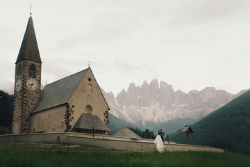 Fototapeta na wymiar Kissing wedding couple stands before stone church in mountains