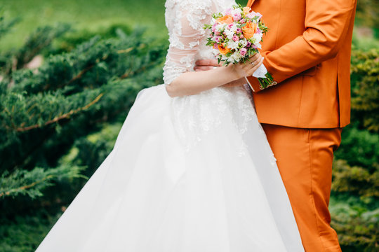 Cropped shot of young newlyweds pose against green nature background. Bride in white dress holds wedding bouquet stands next to groom in orange suit. People, wedding and relationships concept