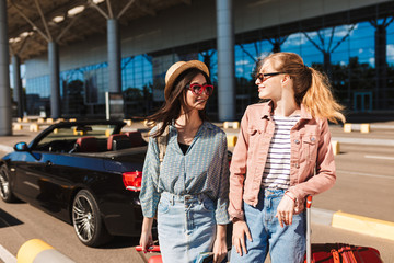 Pretty girls in sunglasses happily looking at each other with cabriolet on background