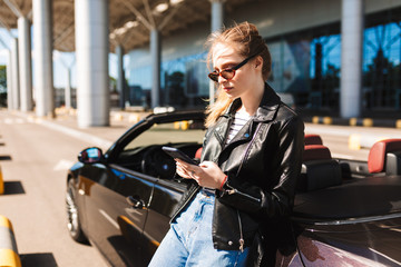 Fototapeta na wymiar Beautiful girl in sunglasses and leather jacket leaning on black cabriolet car while thoughtfully using cellphone with airport on background