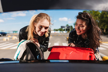 Two cheerful girls happily folding suitcases together into trunk of car in  airport parking zone