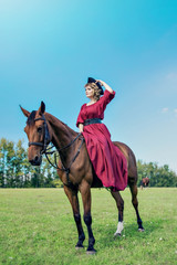 Beautiful girl in a long red dress riding a brown horse against a blue sky.  And she holds her hat so that it will not be blown away by the wind.