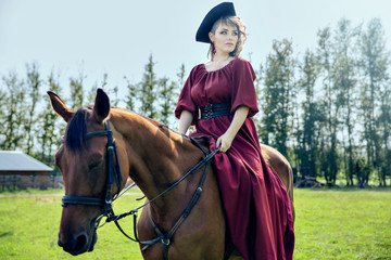 Beautiful girl in a red long red dress and in a black hat with a cocked hat riding a brown horse.