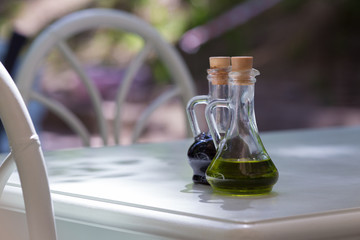 Two jugs, with olive oil and balsamic vinegar on the wooden table. Beside are two chair. On the backgoung is nature.