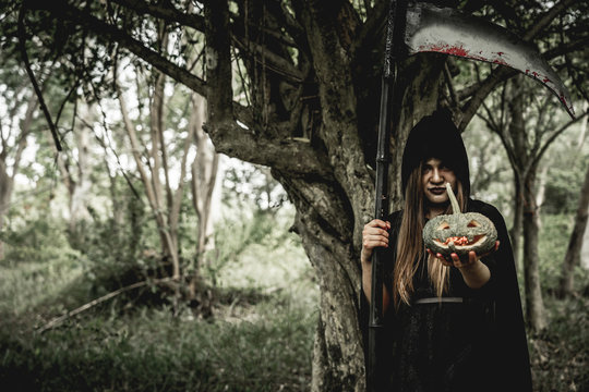 Asian woman dress black as Grim Reaper of death and holding or wielding a large scythe and holding a death pumpkin in Halloween festival. Halloween concept.