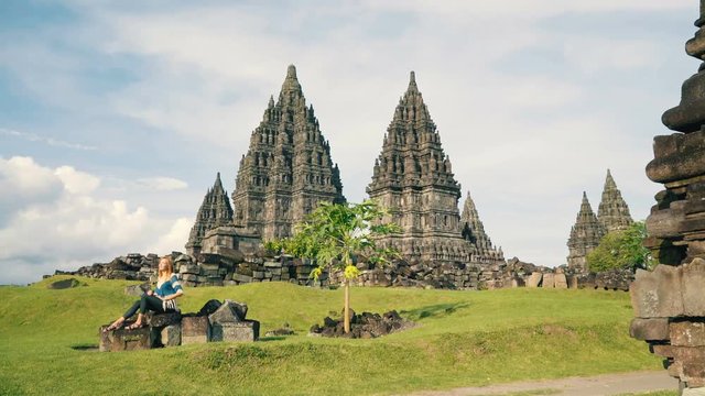 Young caucasian woman sitting on stones on green lawn in front of Prambanan temple on sunny day in Jogjakarta, Indonesia, slide left