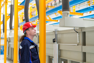 Worker in a factory wearing blue working suit and red helmet looking up in machines in the background