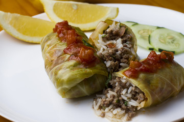 Cigar of cabbage served on white plate accompanied by slices of cucumber, sicilian lemon and tomato on yellow wooden table