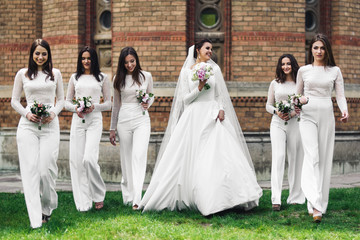 Beautiful bride and bridesmaids with wedding bouquets pose on the green lawn - obrazy, fototapety, plakaty