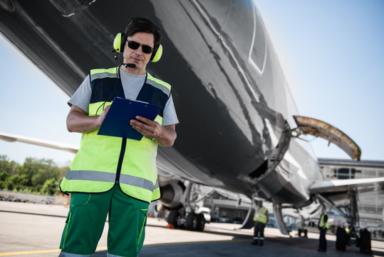 Active teamwork. Low angle portrait of serene man in sunglasses writing on clipboard. Colleagues, plane with open cargo door in the background