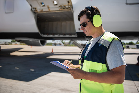 Important data. Side view of serene man in sunglasses and headphones filling out documents. Plane with open cargo door in the background