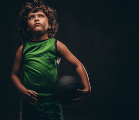 Little basketball player in sportswear holding ball in a studio. Isolated on the dark textured...