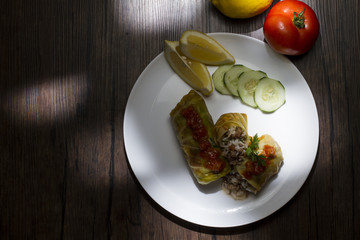 Cigar of cabbage served on white plate accompanied by slices of cucumber, sicilian lemon and tomato on rustic wooden table