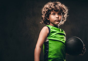 Little basketball player in sportswear holding ball in a studio. Isolated on the dark textured...