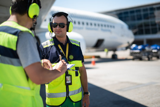 Right here. Smiling man in headphones pointing at clipboard while partner looking at writing data. Passenger plane on blurred background