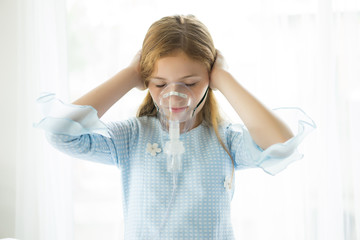 Little girl is sick using oxygen mask on her face whilst his hands covering ears not listening at...