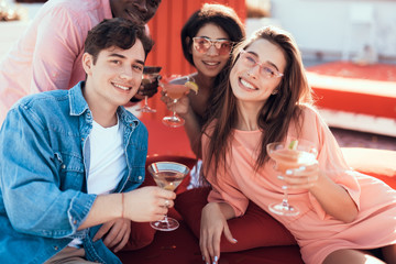 Satisfied males and beaming women drinking alcohol cocktails while looking at camera. Cheerful comrades relaxing together concept