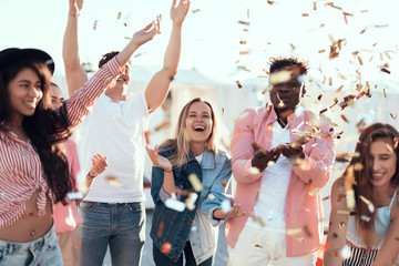 Outgoing males and cheerful women throwing confetti having fun outdoor. Positive comrades spending...