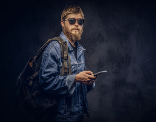 Bearded hipster guy wearing sunglasses and backpack dressed in jeans jacket holds a tablet computer on a dark background.