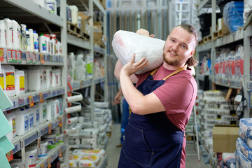 Young bearded salesman carries a pack of plaster on his shoulder in counstruction store