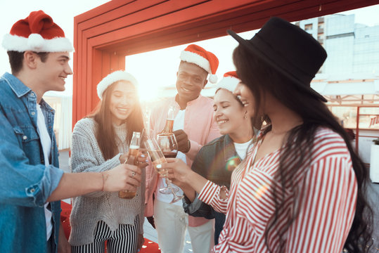 Positive males and cheerful ladies speaking while tasting alcohol liquid. They celebrating new year while wearing red hats